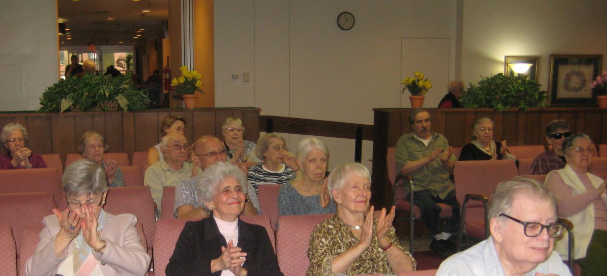 Group of senior adults sitting in rows of seats, some clapping, in a well-lit indoor setting.