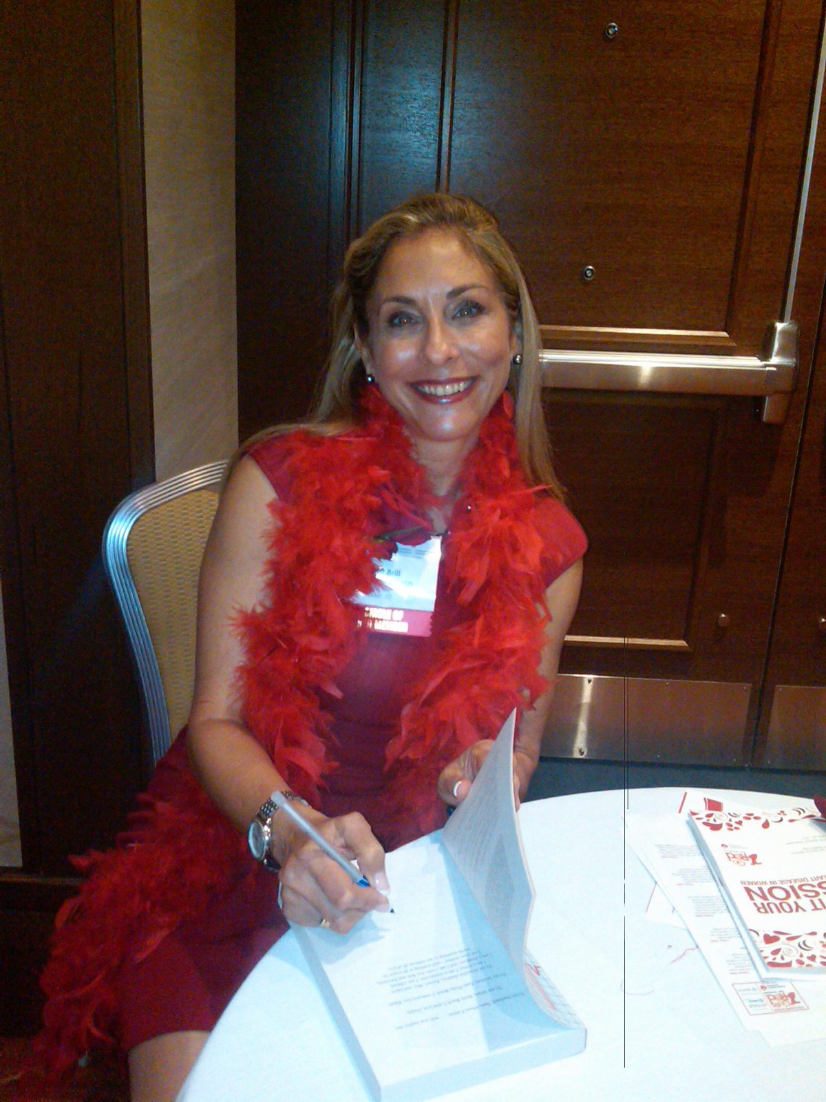 A woman wearing a red dress and a feather boa smiles as she signs a document at a table.